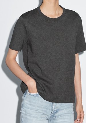 Person wearing a dark gray short-sleeve shirt with light blue jeans, one hand in pocket, standing against a plain white background.