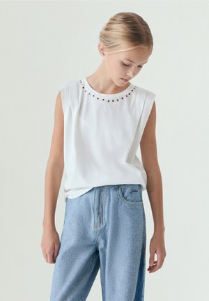 Fille portant un haut blanc sans manches avec des clous métalliques au niveau du col et un jean bleu clair taille haute, regardant vers le bas avec les mains détendues sur les côtés.