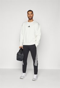 Balts džemperis ar melnu logotipu, apvienots ar melnām šaurām biksēm ar baltām sānu svītrām. Pelēki sporta apavi un melna soma.