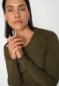 Maslinasto zeleni rebrasti duži rukav majica s okruglim izrezom i nabranim manžetama, u kombinaciji s prirodnim tonovima kože i rukama nježno spletenim.