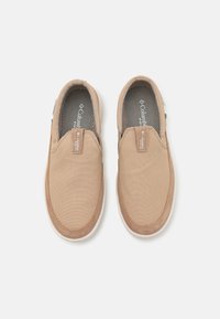 Beżowe płótno buty typu slip-on z teksturowaną górą, kontrastującymi brązowymi akcentami, elastycznymi bocznymi panelami i białą gumową podeszwą.