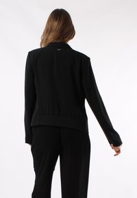 Blazer nero con un design aderente, spalle sagomate e orlo posteriore arricciato. Tessuto liscio e maniche lunghe, senza chiusure visibili.