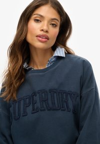 Mujer con cabello largo y ondulado que lleva un sudadera azul marino de Superdry sobre una camisa de cuello a rayas azul y blanca, frente a un fondo liso.