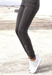 Dunkelgraue Sportleggings mit einem körperbetonten Design, versehen mit einem weißen gestickten Logo an der Seite. Kombiniert mit weißen Turnschuhen.