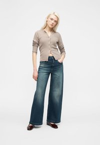 Jeune femme aux longs cheveux blonds portant un cardigan beige boutonné, un jean bleu pantalon large et des chaussures marron foncé, debout avec une main dans la poche.
