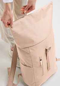 Beige rugzak met een gestructureerde buitenkant, bovenste ritssluiting en zijzakken. Verstelbare schouderbanden en een strak, minimalistisch ontwerp.