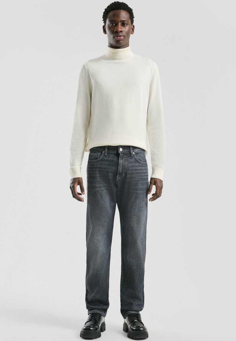 Mężczyzna stoi w białym swetrze z golfem, wyblakłych czarnych dżinsach i czarnych masywnych butach, na jasnym, jednolitym tle.