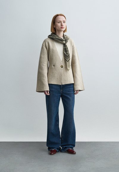 Μπεζ σακάκι από μείγμα μάλλινου υφάσματος με βαθύ ντεκολτέ σε σχήμα V και διπλό κούμπωμα με κουμπιά, σε συνδυασμό με φαρδιές τζιν παντελόνες μπλε χρώματος και εμπριμέ ελιάς πράσινο φουλάρι.