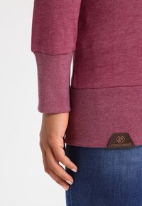 Mano rilassata accanto al corpo, indossando un maglione a maniche a coste color mauve e jeans in denim blu, etichetta del marchio "ragwear" visibile all'orlo del maglione.