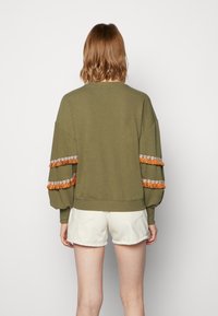 Sudadera verde oliva con hombros caídos y puños elásticos, con detalles de flecos naranjas y bordados en las mangas.