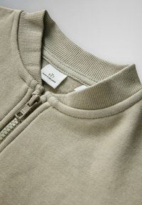 Beige kledingstuk met ritssluiting, geribde kraag en zichtbaar etiket met de tekst "THE SET Made in Sri Lanka."