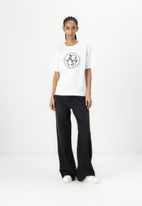 White cotton t-shirt with a round black graphic design, paired with loose-fitting black jeans and white sneakers.