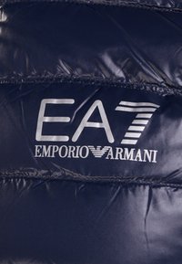 Tmavě modrá polstrovaná látka s bílým logem "EA7 Emporio Armani" potištěným na povrchu.