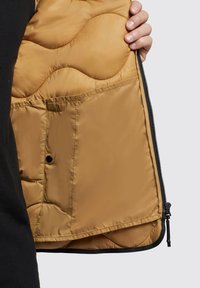 Lightweight beige quilted vest with a smooth texture, featuring a side zip closure and an interior pocket, shown held against a dark garment.
