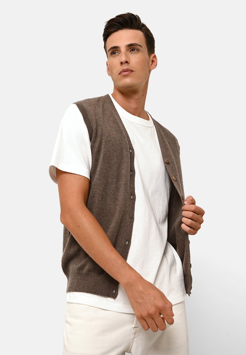 Brun ullkofta med V-hals, knappad framtill och ribbade kanter, över en vit t-shirt, matchad med ljusa byxor.