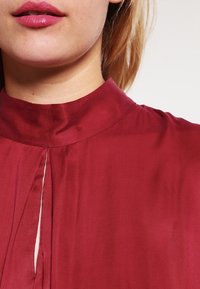 Burgundyröd blus med hög krage och framremsa; slätt, glänsande tyg med en strukturerad yta och subtil glans.