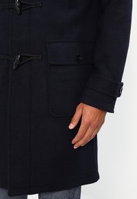 Cappotto di lana blu navy con chiusure a toggles, tasche laterali e vestibilità sartoriale. I polsini presentano dettagli con bottoni. Tessuto testurizzato con una finitura liscia.