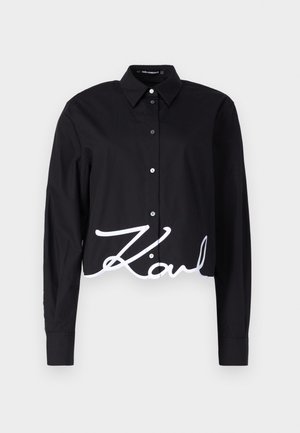 Schwarzes, kurzes Oberteil mit langen Ärmeln, Knopfverschluss und einer weißen, geschwungenen "Karl"-Schrift am Saum. Hergestellt aus glattem Stoff.