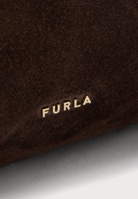 Donkerbruine suède oppervlak met kleine gouden metalen letters die "FURLA" spellen, gecentreerd nabij de onderrand.