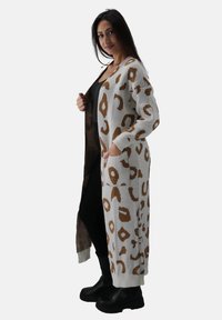 Cárdigan largo de estampado de leopardo en color crema y marrón con textura de punto, con frente abierto, bolsillos y puños acanalados. Usado sobre un atuendo negro.