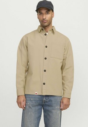 Beige Knopfjacke mit linker Brusttasche, schwarzen Knöpfen und einem Etikett mit rotem Logo, getragen mit blauen Jeans.