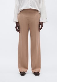 Μπεζ παντελόνι με φαρδύ κόψιμο και ομαλή υφή, με ραμμένη σχεδίαση, μπροστινές τσέπες και διακριτικές ραφές, σε συνδυασμό με λευκό πουκάμισο.