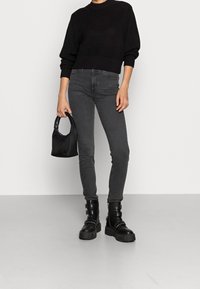 Czarny sweter z dzianiny, szare obcisłe jeansy i masywne czarne botki z klamrami. Szara torebka trzymana w ręce, o minimalistycznym wzornictwie i fakturze.