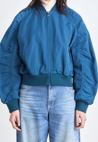 Teal bomberjacka med dragkedja fram, ribbade ärmslut och nederkant samt volangärmar. Matchas med ljusblå denimjeans.