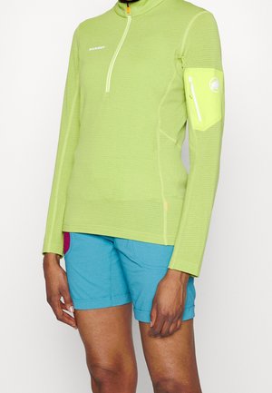 Persoon draagt limegroene top met lange mouwen en ritssluiting op borst en arm, gecombineerd met blauwe short, toont bovenlichaam en armen.