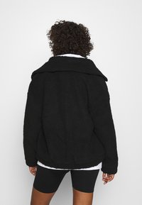 Persoon met krullend haar draagt een zwarte fluffy jas, een wit shirt eronder en zwarte fietsbroek, met de rug naar de camera gericht.