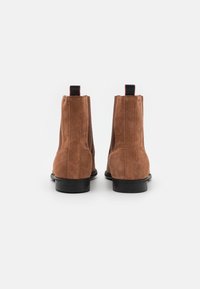 Bottines en daim marron au design élégant, avec des panneaux latéraux élastiques, des semelles en caoutchouc noir et des languettes de talon contrastantes rouges et noires.