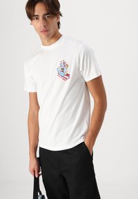 Weißes Baumwoll-T-Shirt mit einem farbenfrohen grafischen Design auf der linken Brust und einem Handmotiv. Der Model steht mit einer Hand in der Tasche und trägt schwarze Hosen.
