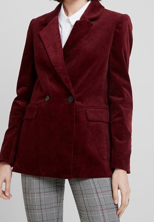 Person wearing a burgundy corduroy blazer with black buttons, white collared shirt, and black-and-white checkered pants, standing against gray background.