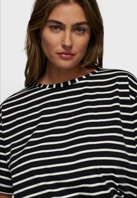 Mujer de cabello castaño claro y ojos azules que lleva una camisa de rayas horizontales en blanco y negro, mirando hacia adelante contra un fondo liso.