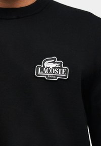 Черна плетена блуза с черно-бял логотип Lacoste Paris, изобразяващ крокодил на гърдите.