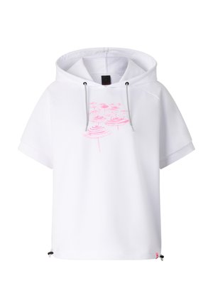 Kobieta o długich ciemnych włosach, ubrana w białą bluzę z kapturem z krótkim rękawem z różowym wzorem, jasnoszarą spódnicę do kolan oraz beżowe trampki z różowymi akcentami.