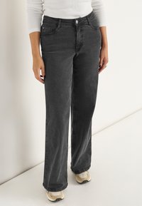 Breda svarta jeans i denim med knappstängning fram, sidofickor och en ren finish vid fållen, båda tillsammans med beige sneakers.