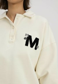 Crèmekleurig polo-shirt met een kraag, met een zwart geborduurd M-logo en wereldboldetail aan de linkerkant.