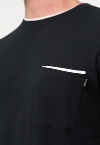 Κοντινό πλάνο σε μαύρο υφασμάτινο πουκάμισο με υφή που δείχνει μια τσέπη στο στήθος με λευκό περίγραμμα και μια μικρή μαύρη υφασμάτινη ετικέτα στο πλάι.