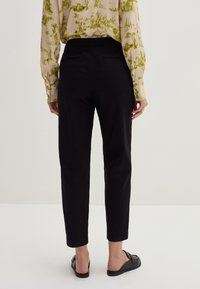 Pantaloni neri a sigaretta con una texture liscia, dotati di una tasca posteriore. Indossati con una blusa chiara a fantasia e sandali neri bassi.