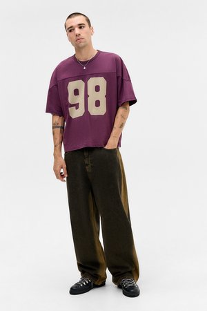 Man with tattoos wearing a purple oversized shirt with number 98 and wide dark pants standing with one hand in pocket.