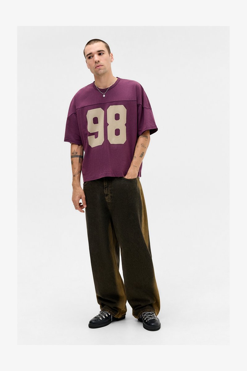 Mand med tatoveringer iført en lilla oversized skjorte med nummer 98 og brede mørke bukser, der står med den ene hånd i lommen.