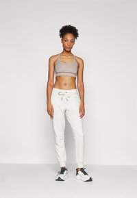 Kvinne som står iført beige sports-bh, hvite sweatpants med snor og hvite sneakers med svarte lisser mot en ensfarget bakgrunn.