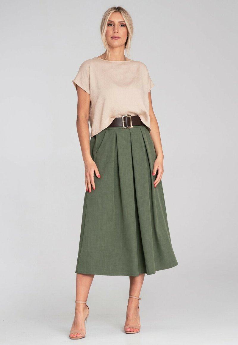 Jasno beżowa bluzka z krótkim rękawem w połączeniu z zieloną, plisowaną spódnicą midi i szerokim brązowym paskiem. Pasek ma dużą srebrną klamrę.