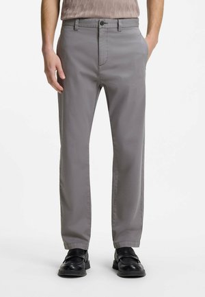 Mann trägt graue gerade geschnittene Hose, schwarze Anzugschuhe und ein helles, gemustertes Hemd, steht mit den Händen in den Taschen.