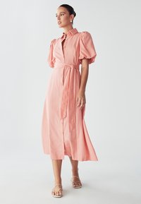 Haljina midi s napuhanim rukavima, dugmadi i svijetlo ružičastim materijalom, s pojasom oko struka. Glatka tekstura, lepršavi silueta i minimalistički dizajn.