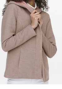 Woman wearing a beige textured hooded jacket over a white top, holding the jacket collar with both hands.