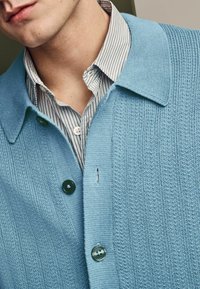 Lysblå strikket cardigan med krave, der har tre knapper. Under den ses en hvid- og blåstribet skjorte delvist.