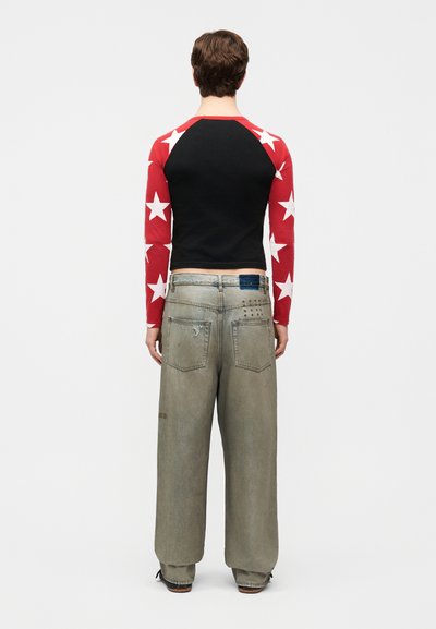 Osoba viditeľná zozadu, oblečená v čiernom tričku s červenými rukávmi so vzorom hviezd a vo voľných, vyblednutých sivých džínsoch s nášivkou na zadnej strane.