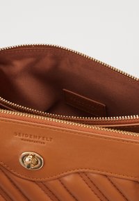 Sisätilat vaaleanruskeassa nahkalaukussa, jossa on pronssinvärinen vetoketju, sileä vuori ja hienovaraisesti kohokuvioitu logo. Vaatimattomat laskostussuunnittelut korostavat laukkujen sisustusta.